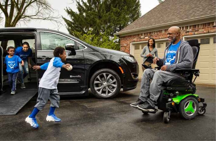 man in wheelchair in his driveway with a wheelchair van and his grandkids running around