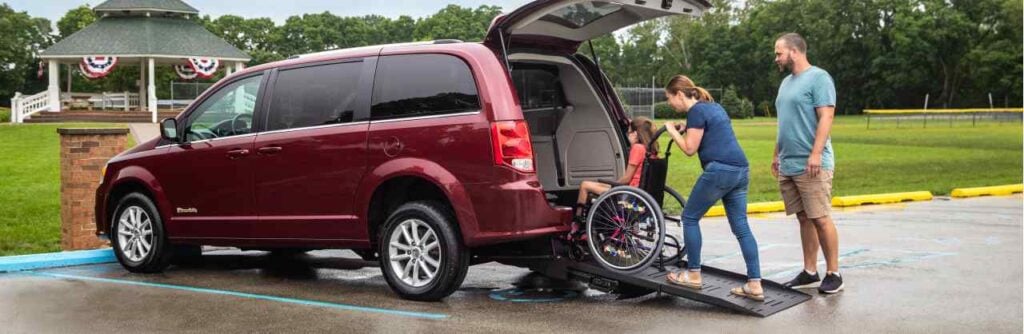 Image of a man, his wife pushing their daughter in a manual wheelchair up the ramp of their red dodge caravan wheelchair van at a park