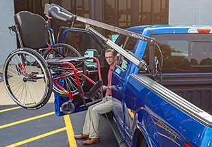 Man operates a lift to load a wheelchair into a blue truck, showcasing mobility solutions at a vehicle dealer.